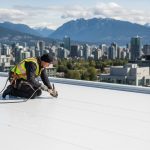 Professional image of a roofer installing TPO membrane on a Vancouver commercial building under cloudy skies with mountain views.