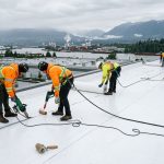 Skilled commercial roofers installing flat TPO roof on warehouse in Delta, British Columbia, with safety gear under cloudy coastal sky