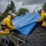 Emergency roof repair team in yellow gear securing blue tarp on damaged suburban roof during Vancouver rainstorm.