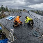 Skilled roofing team in safety gear installing asphalt shingles on residential roof in rainy Vancouver neighborhood, with tools and misty urban backdrop.