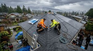 Skilled roofing team in safety gear installing asphalt shingles on residential roof in rainy Vancouver neighborhood, with tools and misty urban backdrop.
