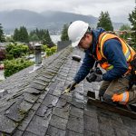 Professional roofer inspecting damaged asphalt shingles on rainy Vancouver residential roof with safety gear and tools, coastal backdrop.